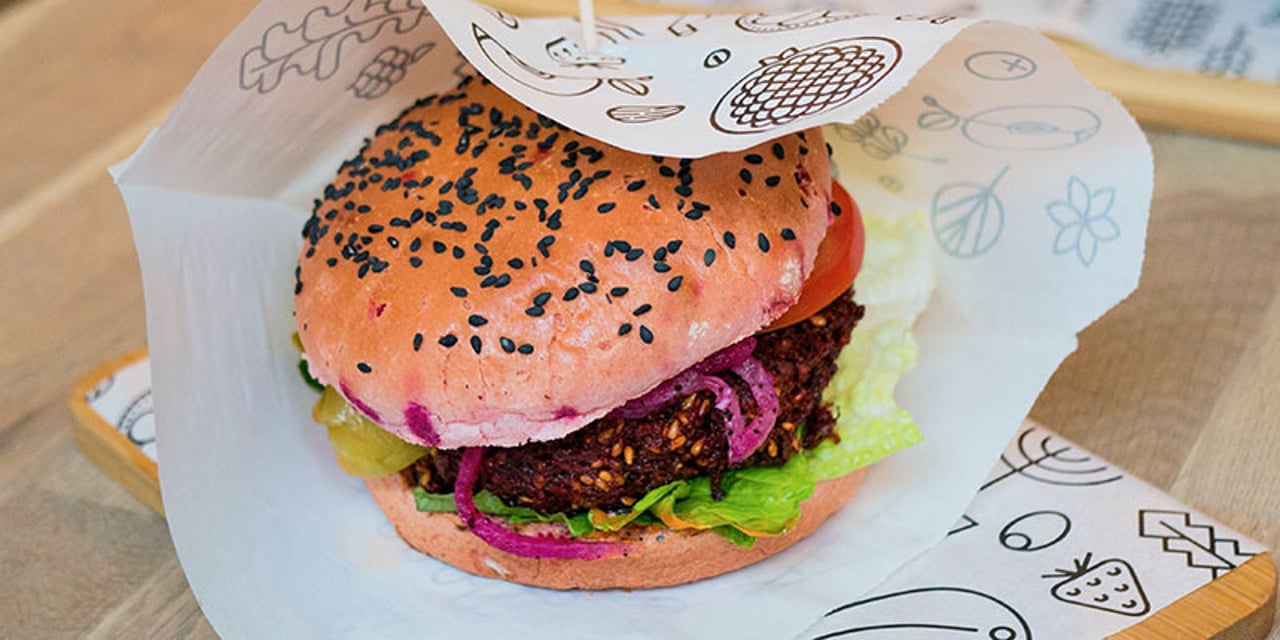 A vegetarian burger in a bun with colourful garnishes, served on white greaseproof paper with black and white designs of vegetables